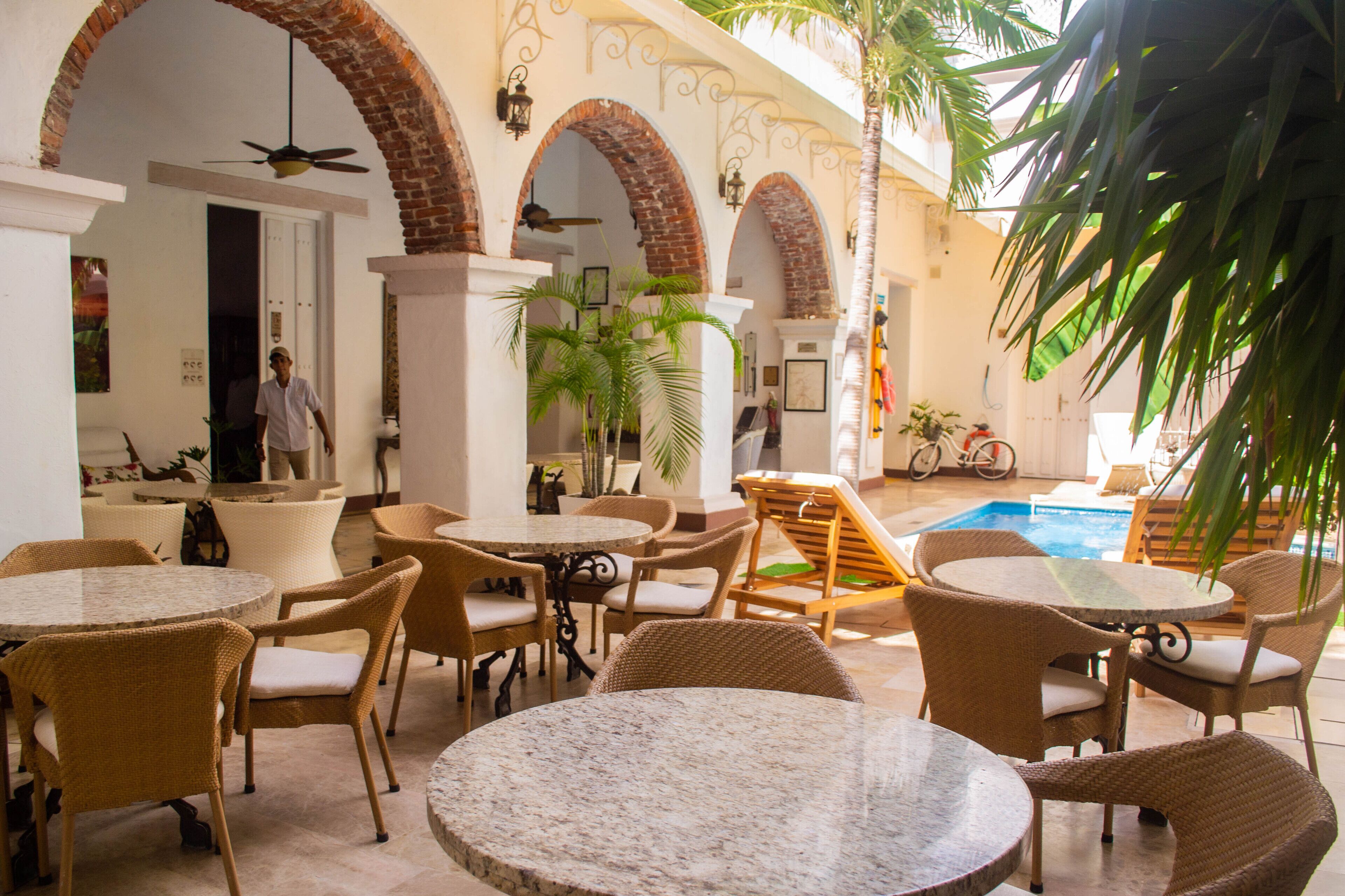 A poolside area with multiple round tables and wicker chairs, surrounded by brick arches and potted plants.