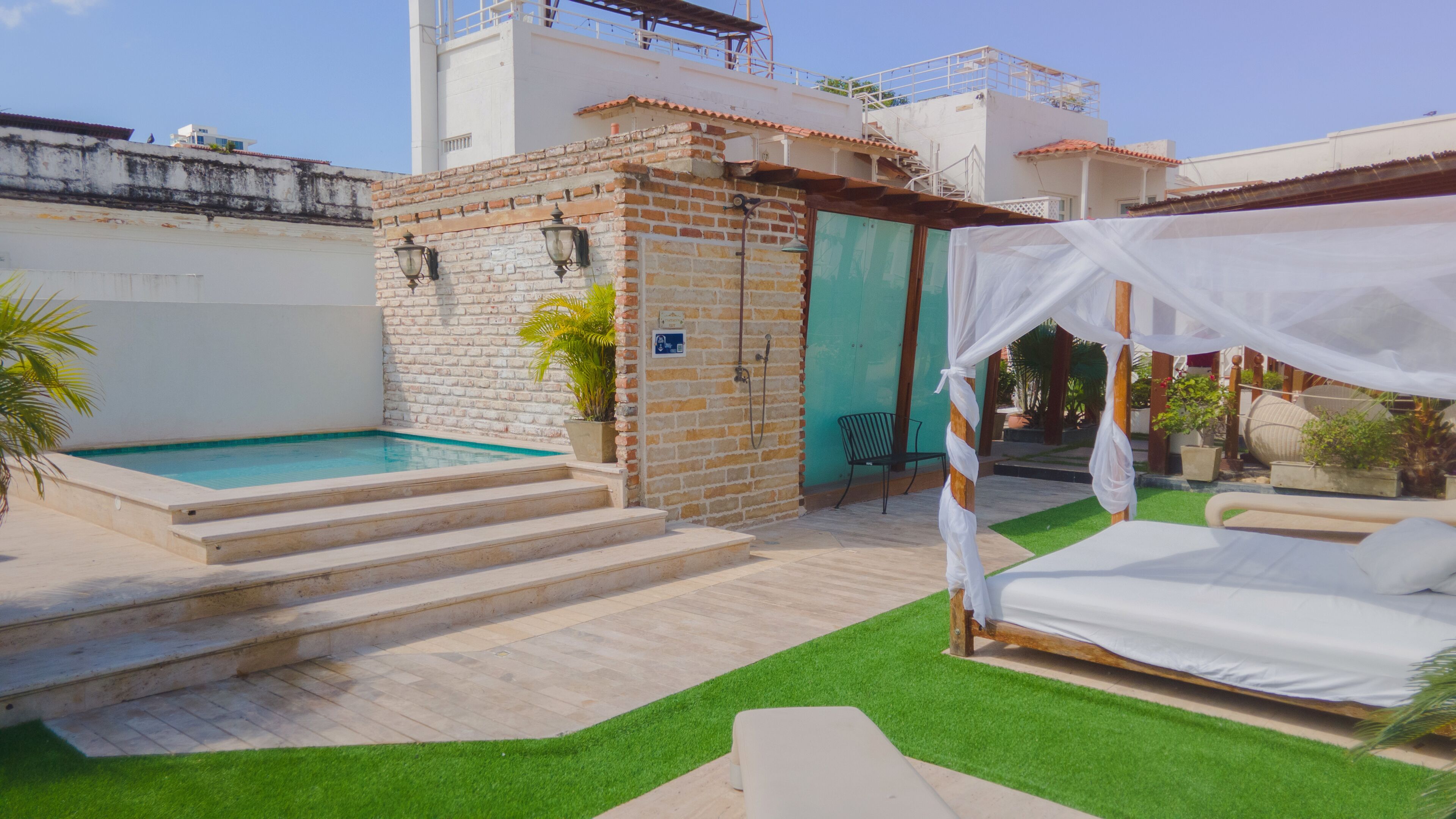 Un área de piscina con escalones que llevan a una puerta, una cama con dosel y un pequeño jardín con césped artificial y plantas en macetas.