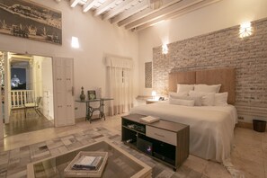 A bedroom with a large bed, a wooden headboard, a brick accent wall, a small table with a lamp, and a glass coffee table. - Hotel Boutique Don Pepe (Santa Marta)
