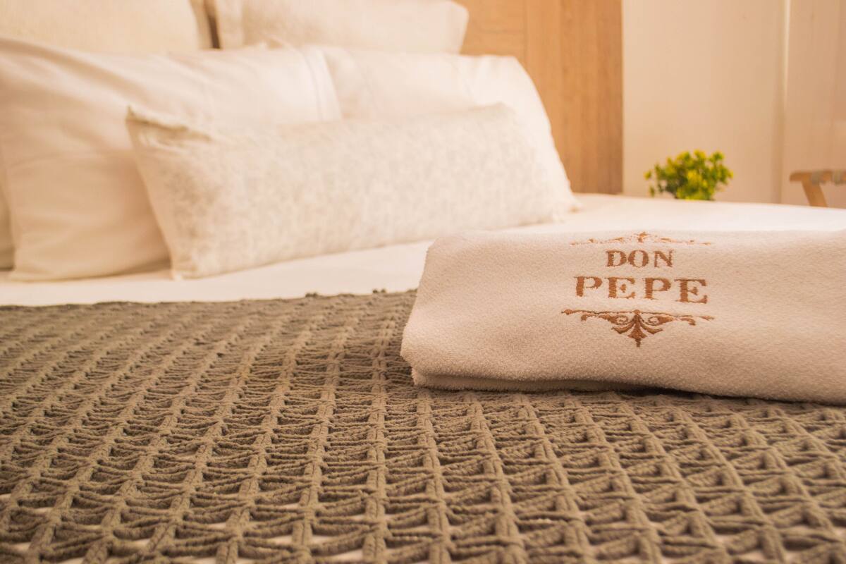 A neatly made bed with a beige blanket and a pillow with "DON PEPE" embroidered on it.