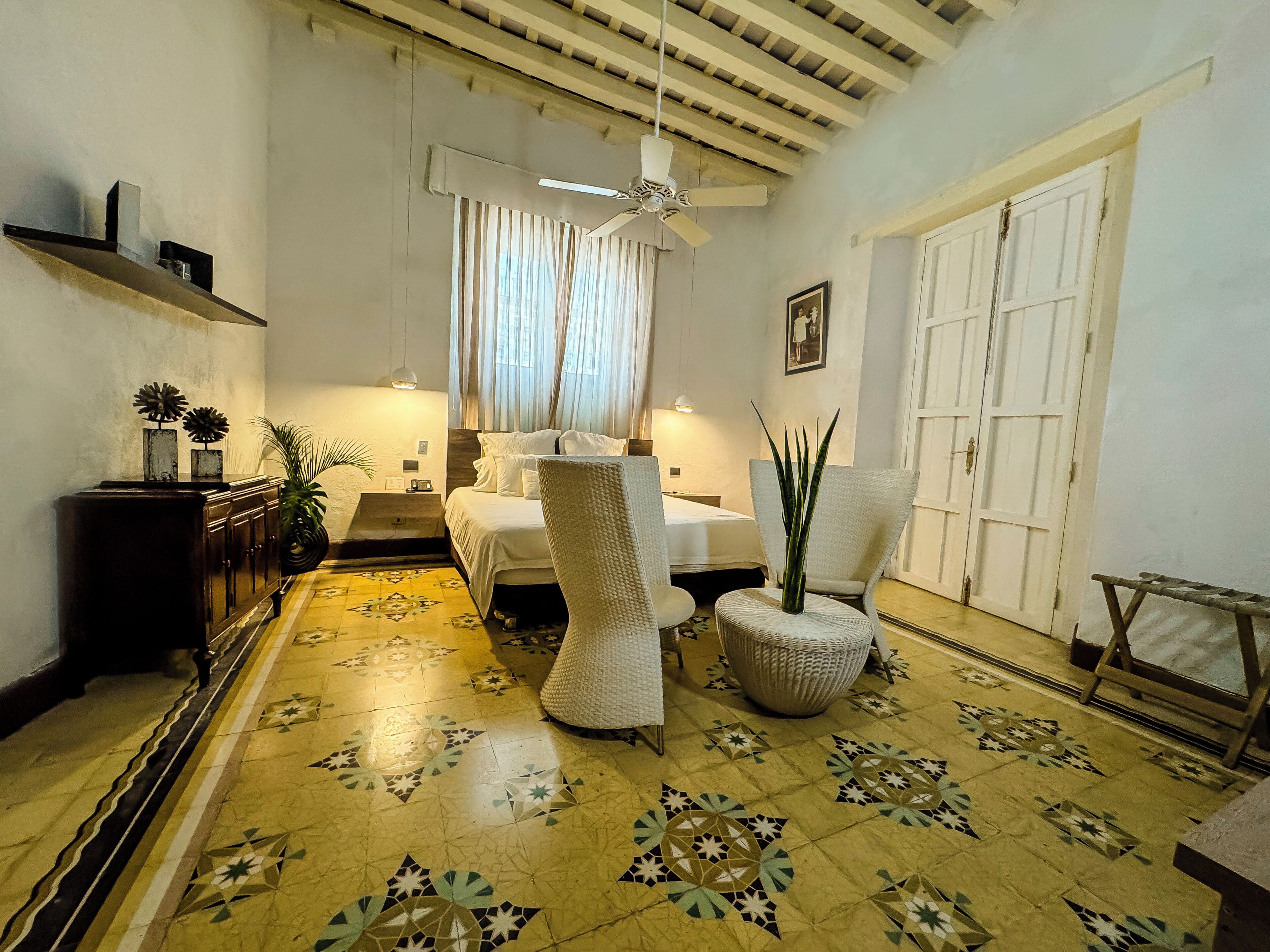 Una habitación bien iluminada con una cama, dos sillones, una mesa, una lámpara de pie y un ventilador de techo.