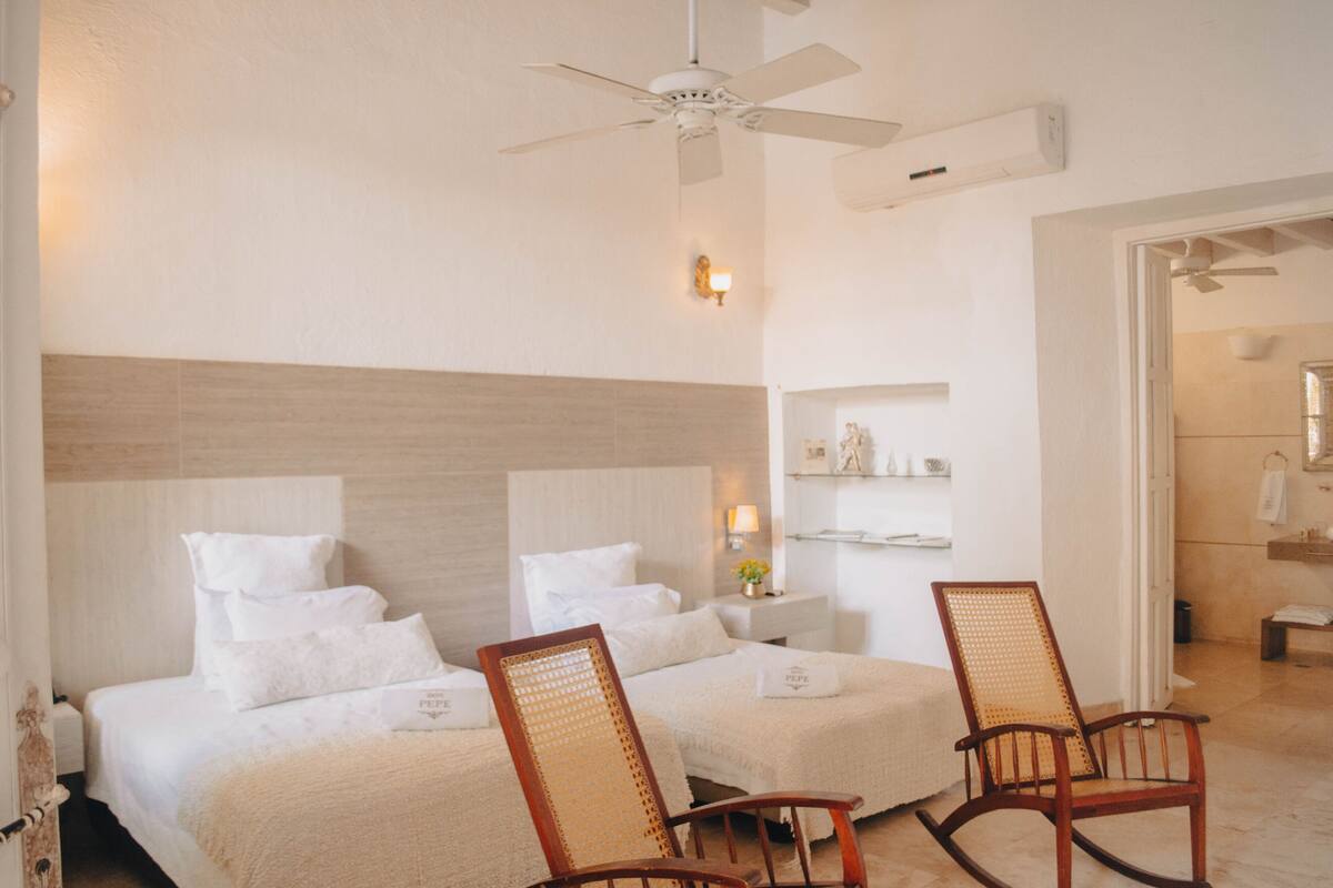 A hotel room with two beds, a ceiling fan, a nightstand with a lamp, and a bathroom visible through an open door.