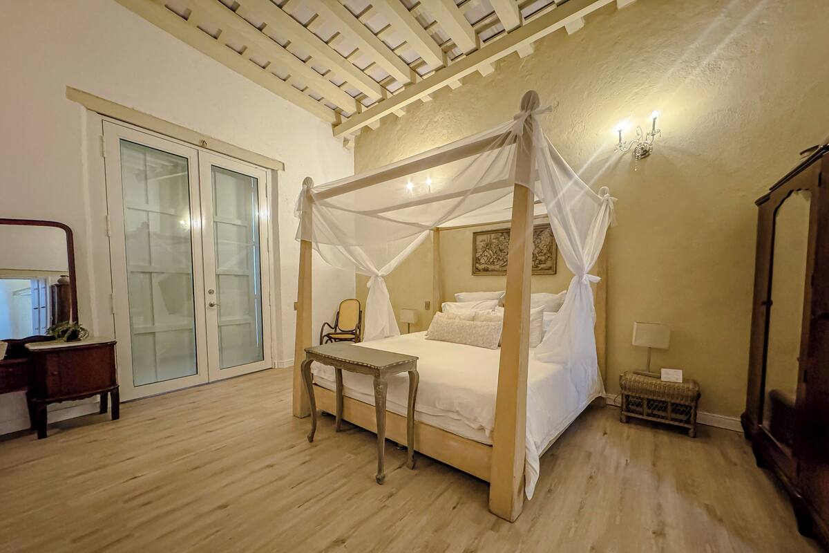 A bedroom with a four-poster bed, a wooden desk, a chair, a wardrobe, and a glass door leading to a balcony.