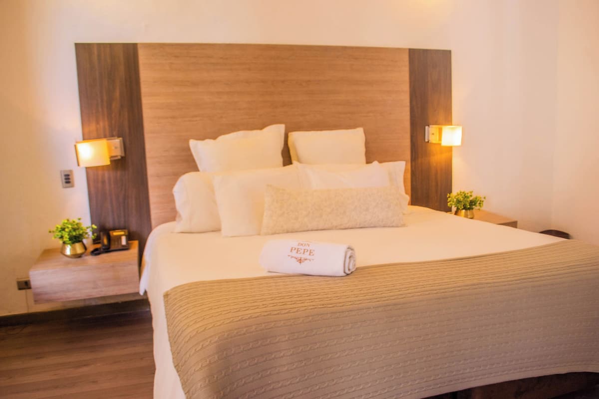 A neatly made bed with a rolled towel, two bedside tables with lamps, and a small plant on each table.
