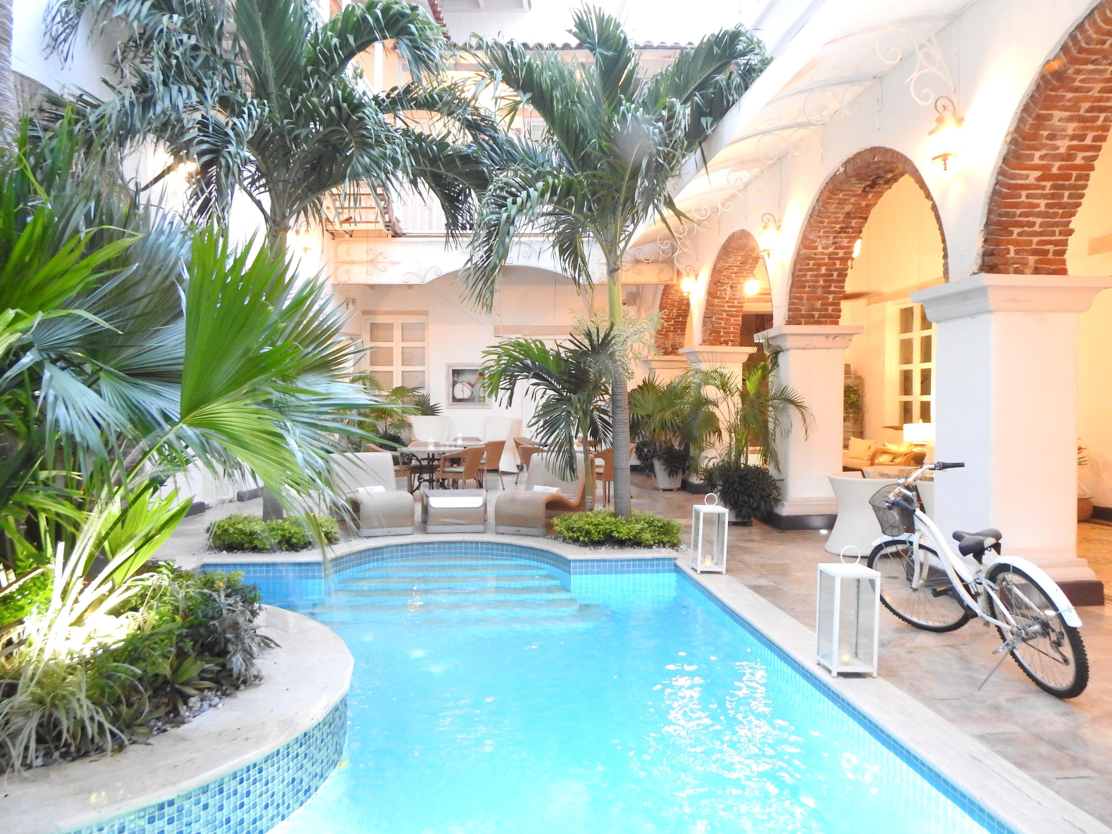 An indoor swimming pool with a tiled edge, surrounded by palm trees and potted plants. There are outdoor seating areas and a bicycle parked nearby.