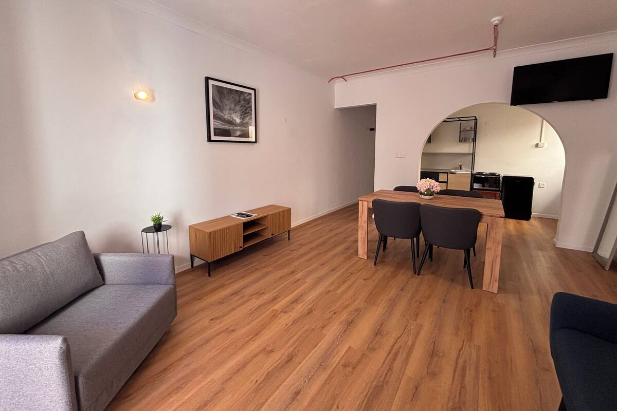 Un salon moderne comprenant un canapé, une table basse en bois, une table à manger avec des chaises, une petite table d’appoint avec une plante et un tableau encadré accroché au mur.