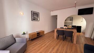 Una sala de estar moderna con un sofá, una mesa de centro de madera, una mesa de comedor con sillas, una mesita auxiliar con una planta y un cuadro en la pared.