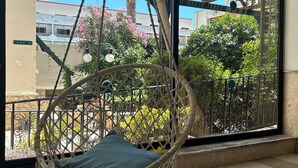 Un balcon spacieux agrémenté d’un fauteuil suspendu, de grandes fenêtres et offrant une vue sur la verdure et les bâtiments environnants.