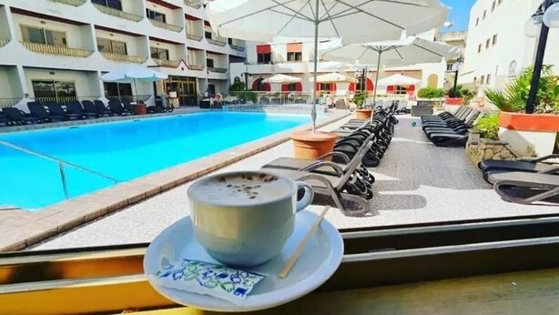 Une vue sur la piscine, avec des chaises longues et des parasols, une tasse de café sur une table, et un bâtiment avec des balcons en arrière-plan.