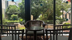 Un espace salon avec une table ronde et des chaises, de grandes fenêtres offrant une vue sur la verdure et les bâtiments, et une structure en forme de pergola.