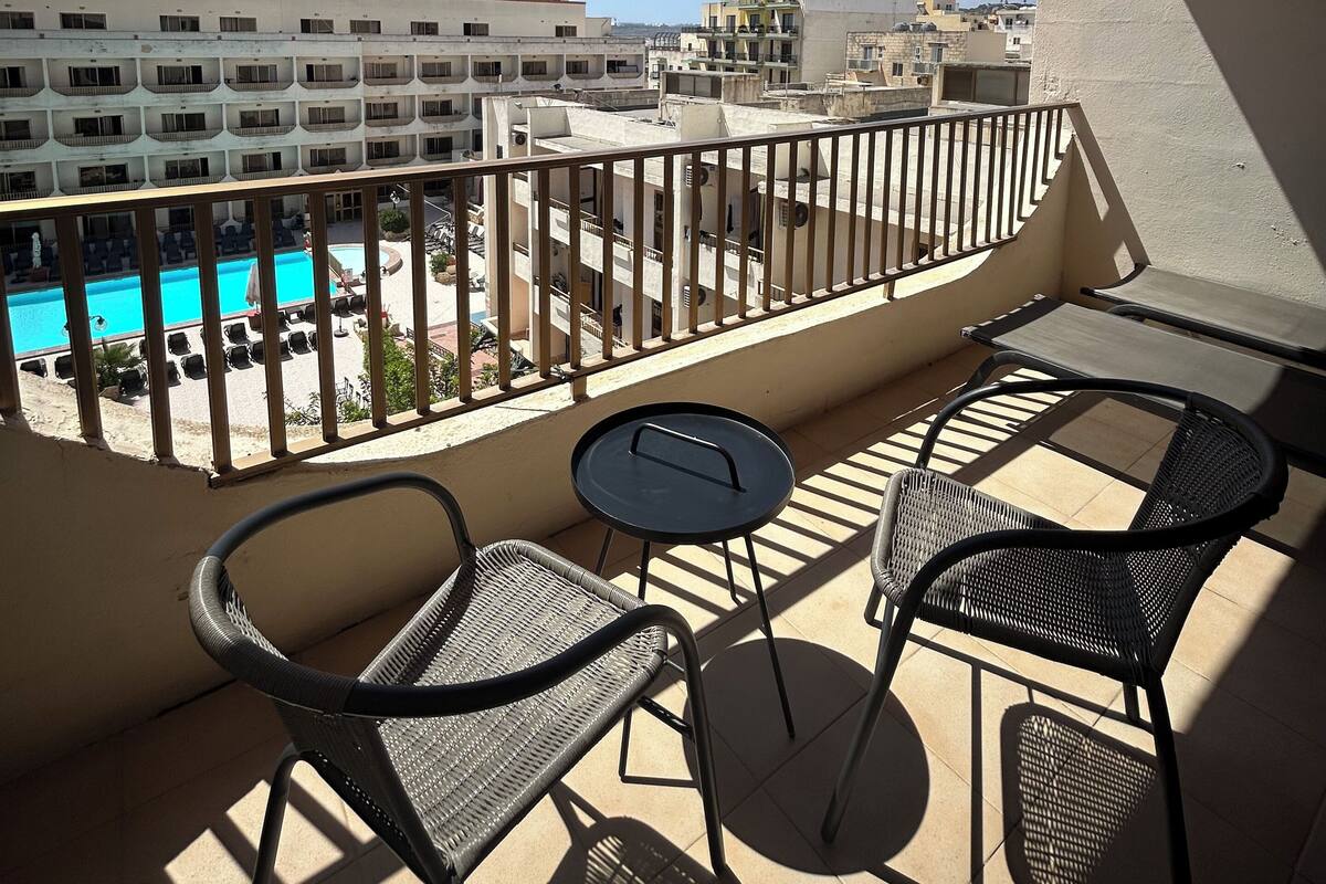 Un balcon avec deux chaises et une petite table, donnant sur une piscine et des immeubles.