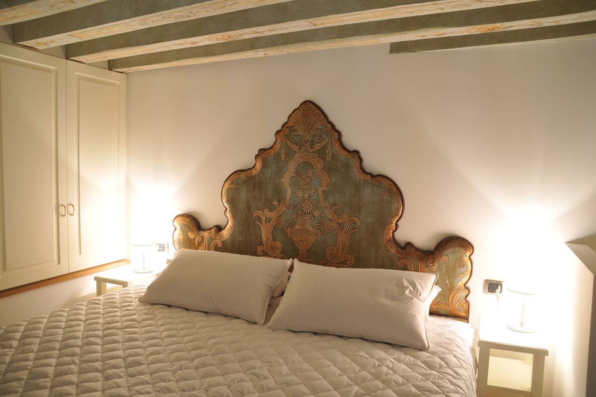 Una cama con colcha acolchada y dos almohadas, cabecero de madera con intrincados adornos, y mesita de noche con una lámpara.