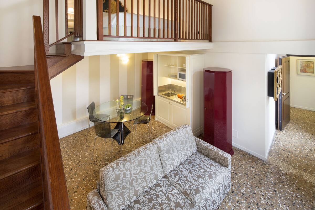 Una casa de dos pisos con una escalera de madera, una mesa de comedor de vidrio, un sofá estampado y una cocina con alacenas y un refrigerador.