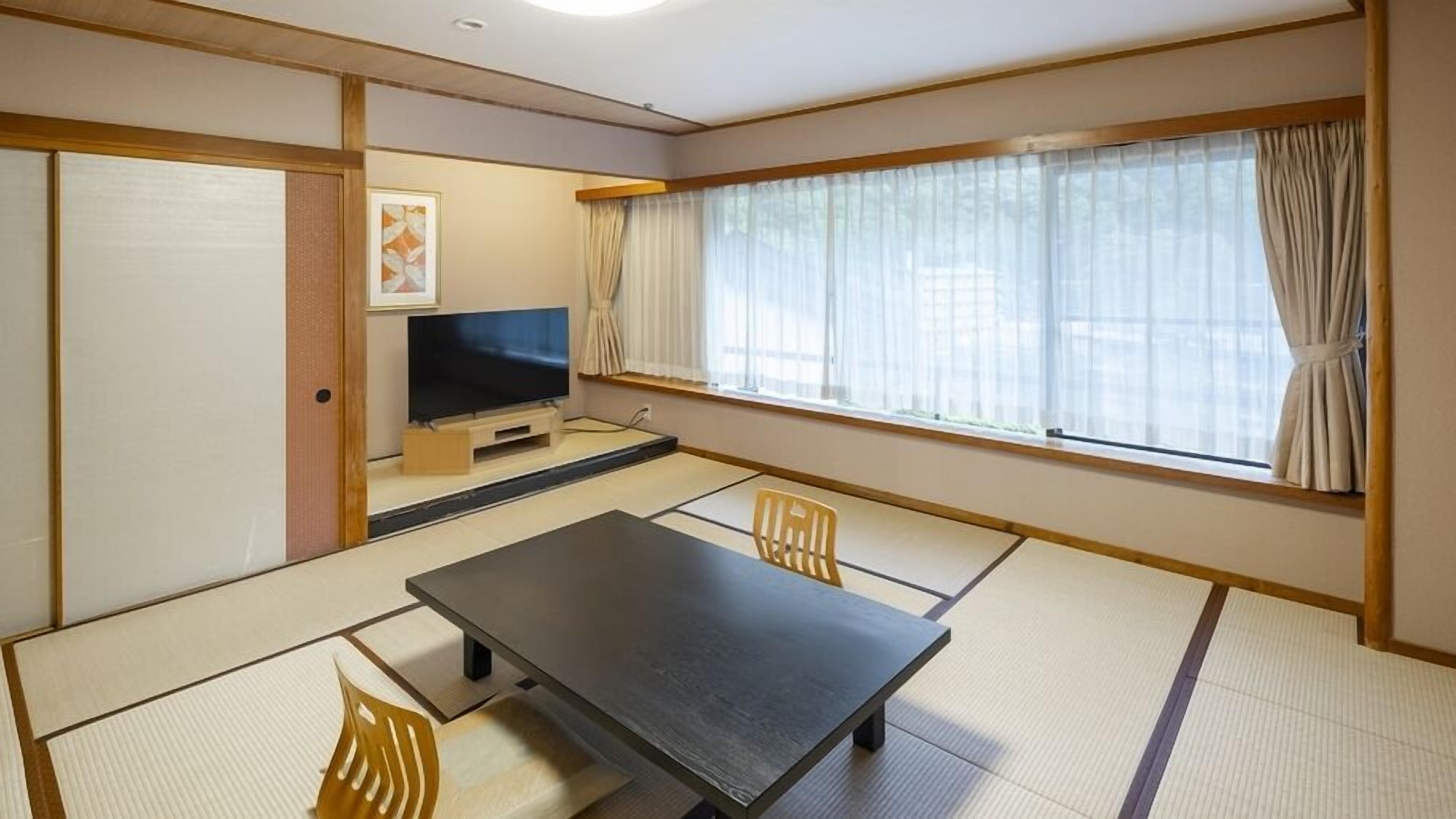 A traditional Japanese room with tatami flooring, a low table, two chairs, a television, and a window with curtains.