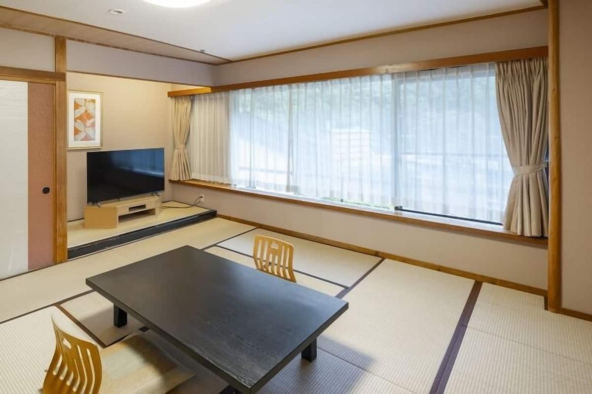 Una habitación tradicional japonesa con suelo de tatami, una mesa baja, dos sillas, un televisor y una ventana con cortinas.
