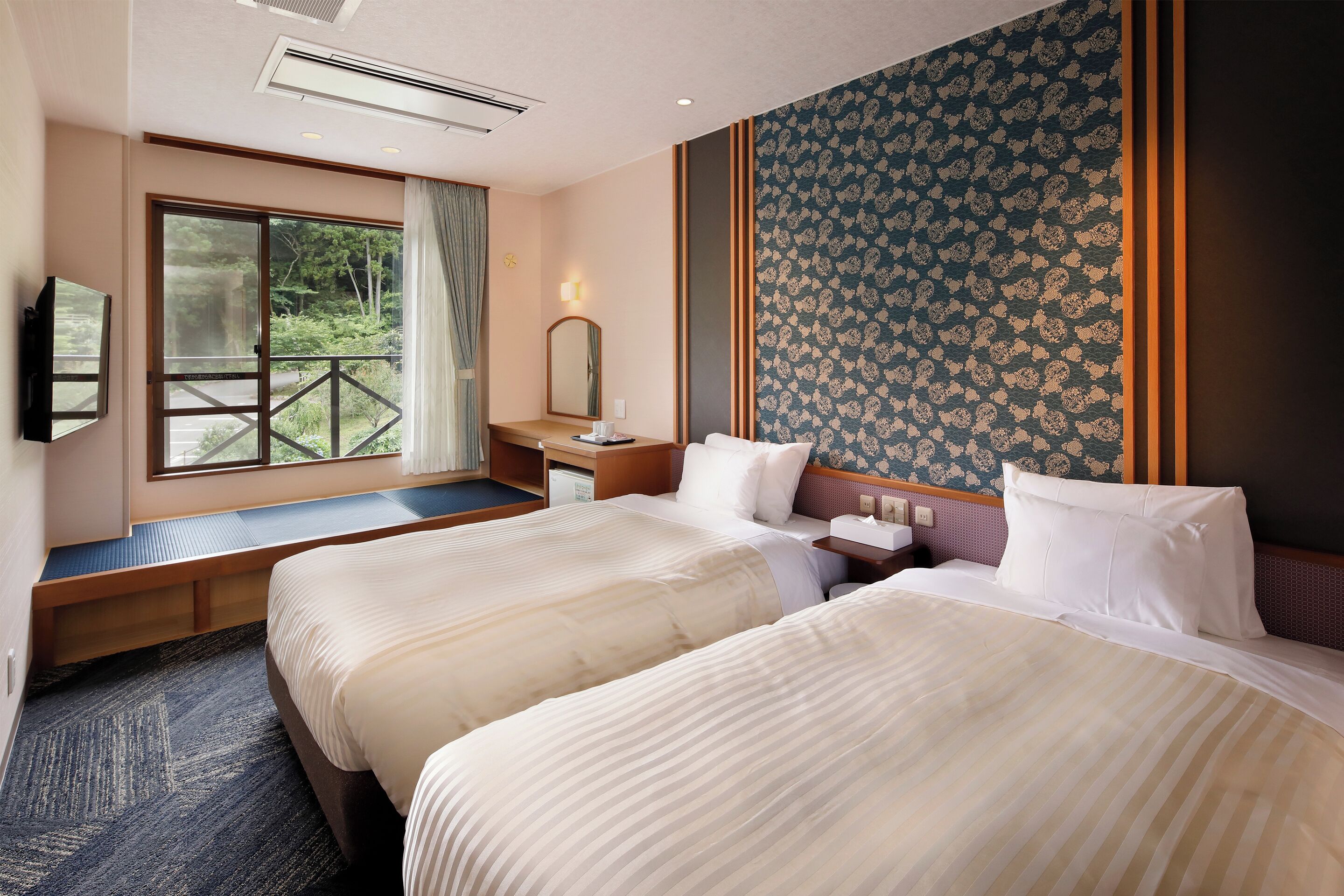 Habitación de hotel con dos camas, un escritorio, una ventana con vistas a zonas verdes y una pared con estampado.