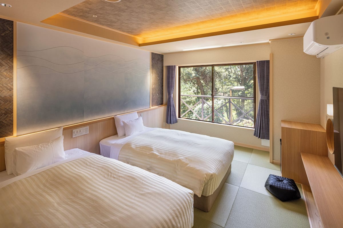 Habitación de hotel con dos camas, un ventanal con cortinas, un banco de madera y un televisor.