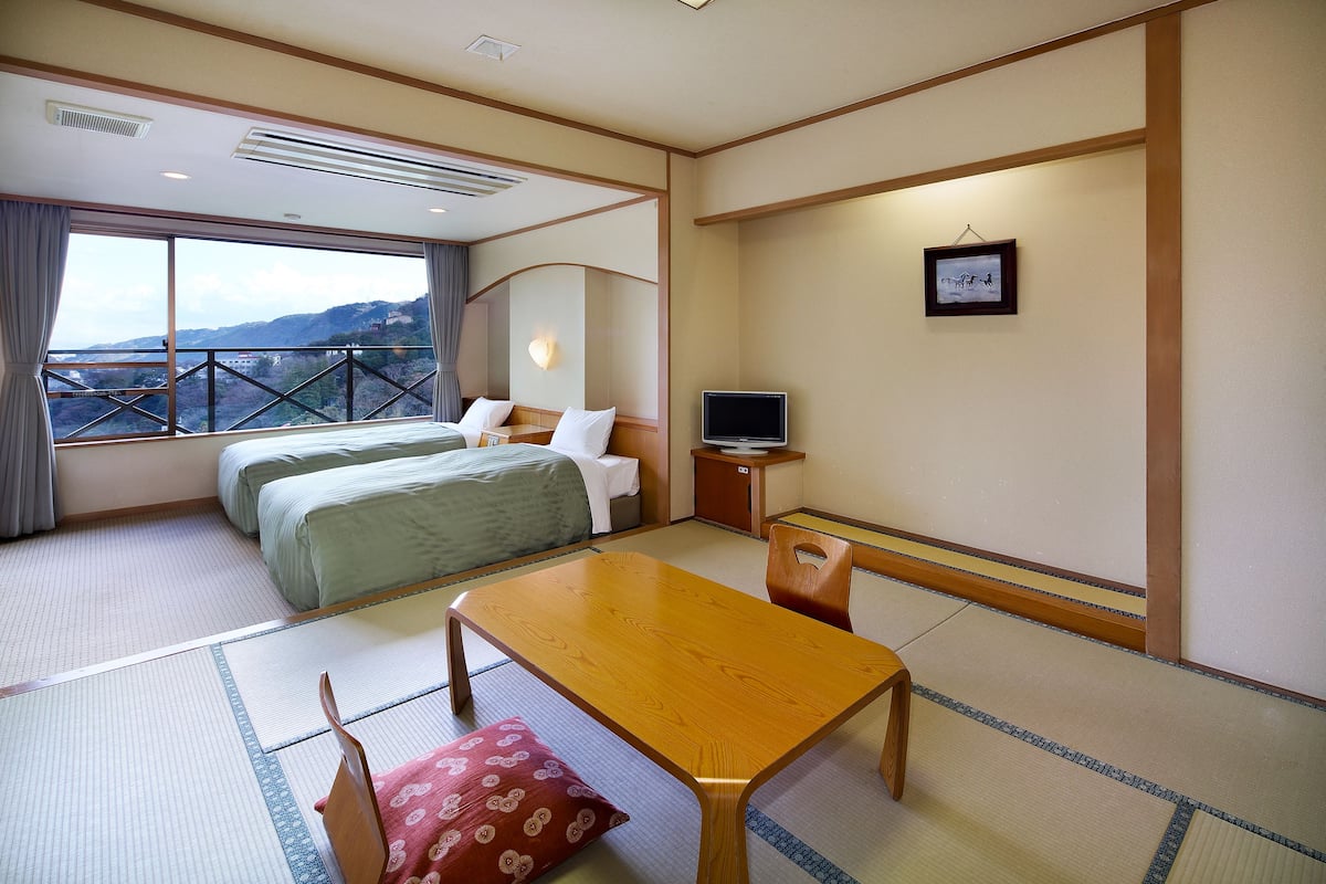 Habitación de hotel con dos camas, una mesa de madera, esteras de tatami, un televisor y vistas a las colinas.