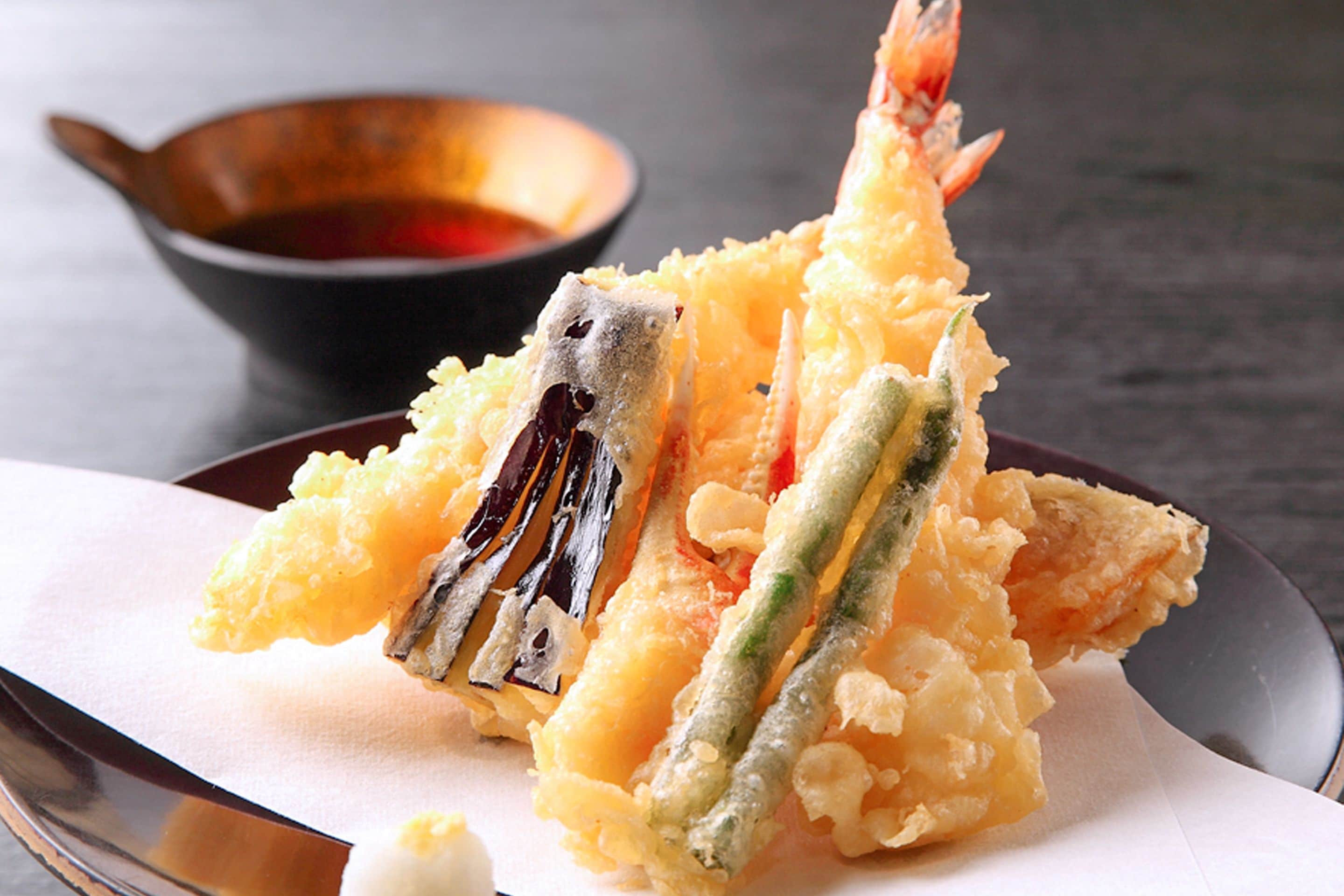 A plate of tempura with shrimp and vegetables, accompanied by a dipping sauce in a small bowl.