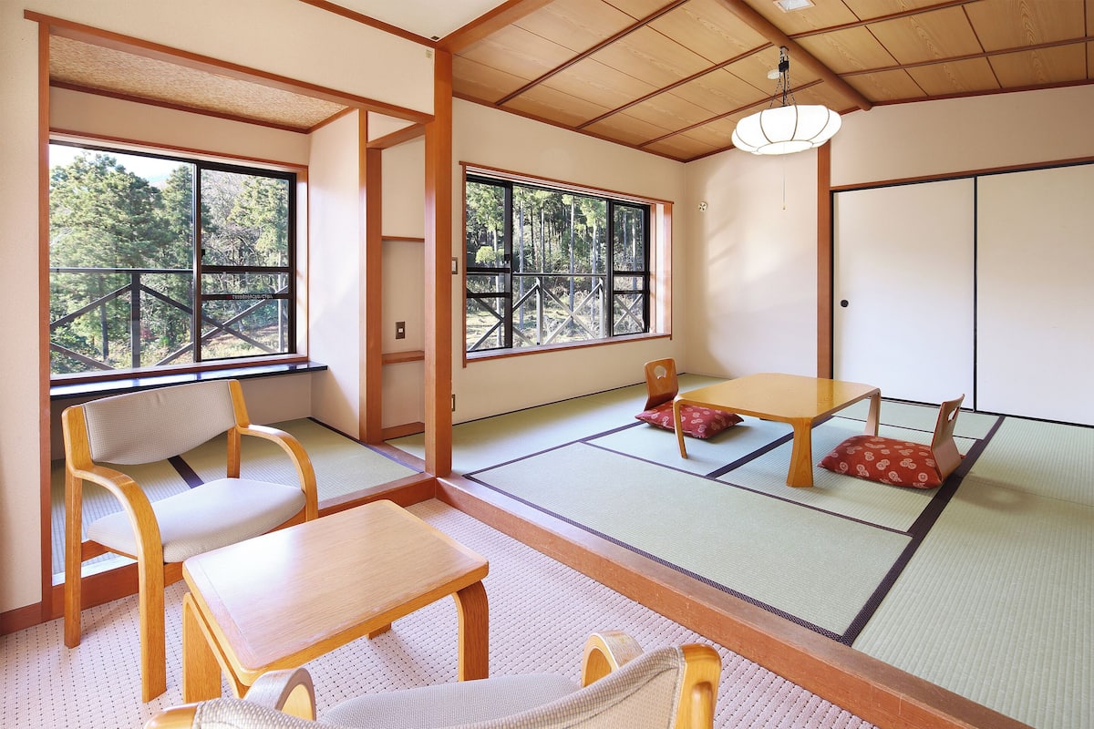Una habitación japonesa tradicional con suelo de tatami, una mesa de madera baja, un banco acolchado y un amplio ventanal con vistas a un bosque.