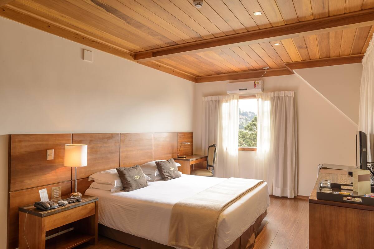 Ein Hotelzimmer mit Holzboden, einem Bett mit weißen Leinen, einem Nachttisch mit Lampe, einem Schreibtisch mit Stuhl und einer Klimaanlage.