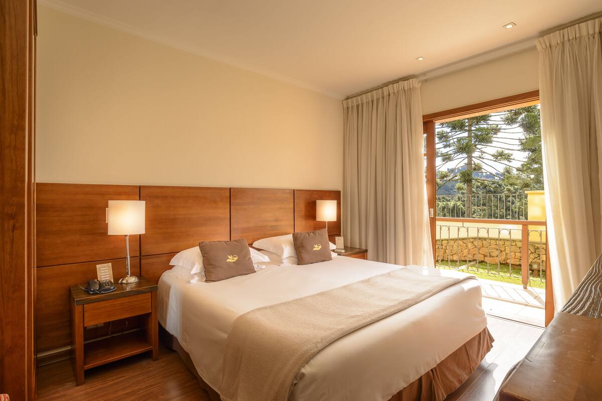 Ein Hotelzimmer mit einem großen Bett, zwei Nachttischen mit Lampen, einem hölzernen Kopfteil und einem Balkon mit Steinmauer und Grünpflanzen.