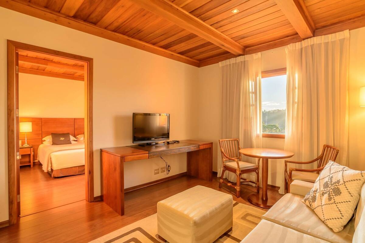 Ein Hotelzimmer mit Holzbalkendecke, einem Bett, einem Fernseher, einem Schreibtisch, einem Stuhl, einem Tisch und Blick ins Freie.
