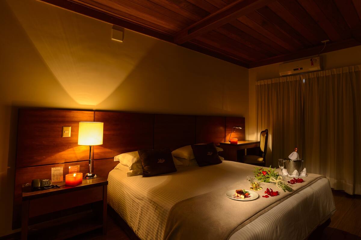 Ein Hotelzimmer mit Holz-Kopfteil, einem Nachttisch mit Lampe sowie einem weiteren Nachttisch mit Telefon und einem mit sauberer Bettdecke bezogenen Bett.
