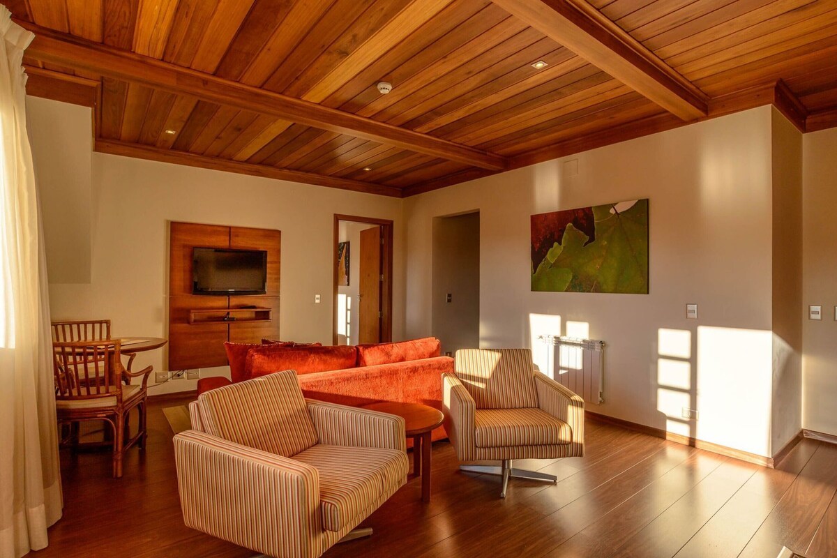A spacious living room with wooden flooring, a large orange sofa, striped armchairs, a wooden ceiling, and a television mounted on the wall.