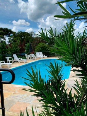 A swimming pool with white lounge chairs and green foliage in the foreground. - Hotel San Lucas (Gramado)