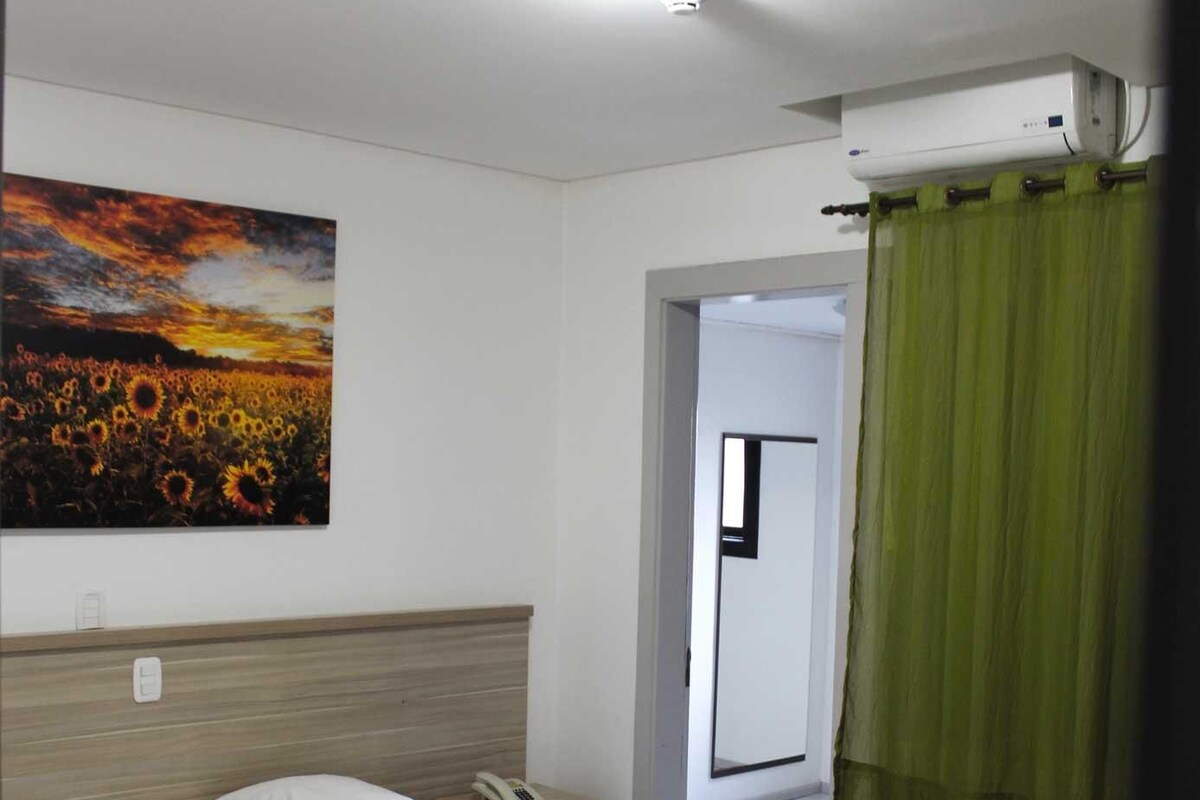 A neatly made bed with a white headboard, a bedside table with a phone, and a painting of sunflowers on the wall.
