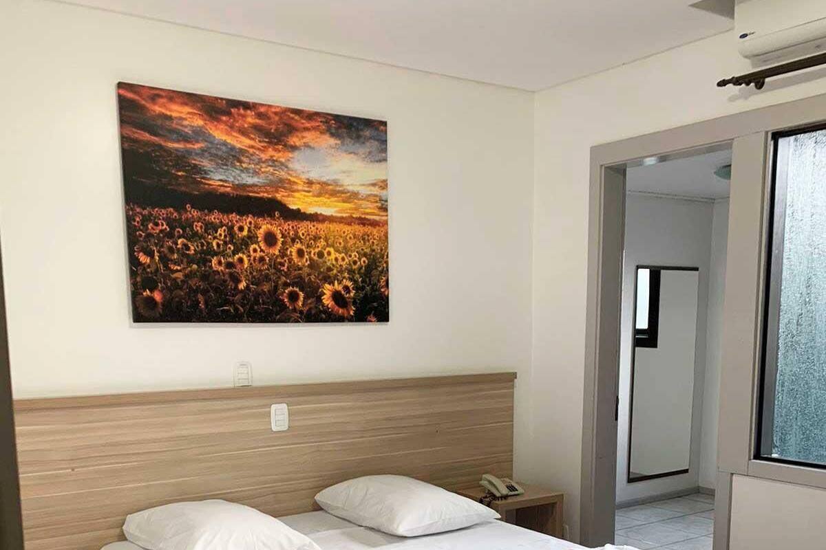 A hotel room with a large bed, a nightstand, a painting of a sunset, and a view through a window.