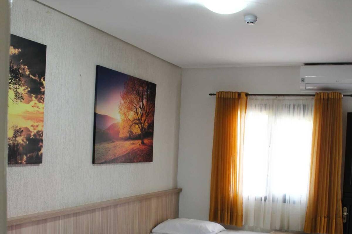 A hotel room with two beds, a window with curtains, and two paintings on the wall.