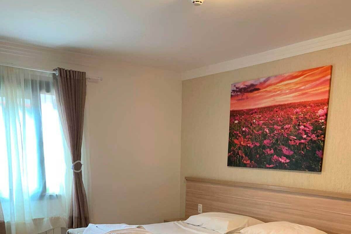 A neatly made bed with white linens and a brown throw blanket, a wooden headboard, and a painting of flowers on the wall.