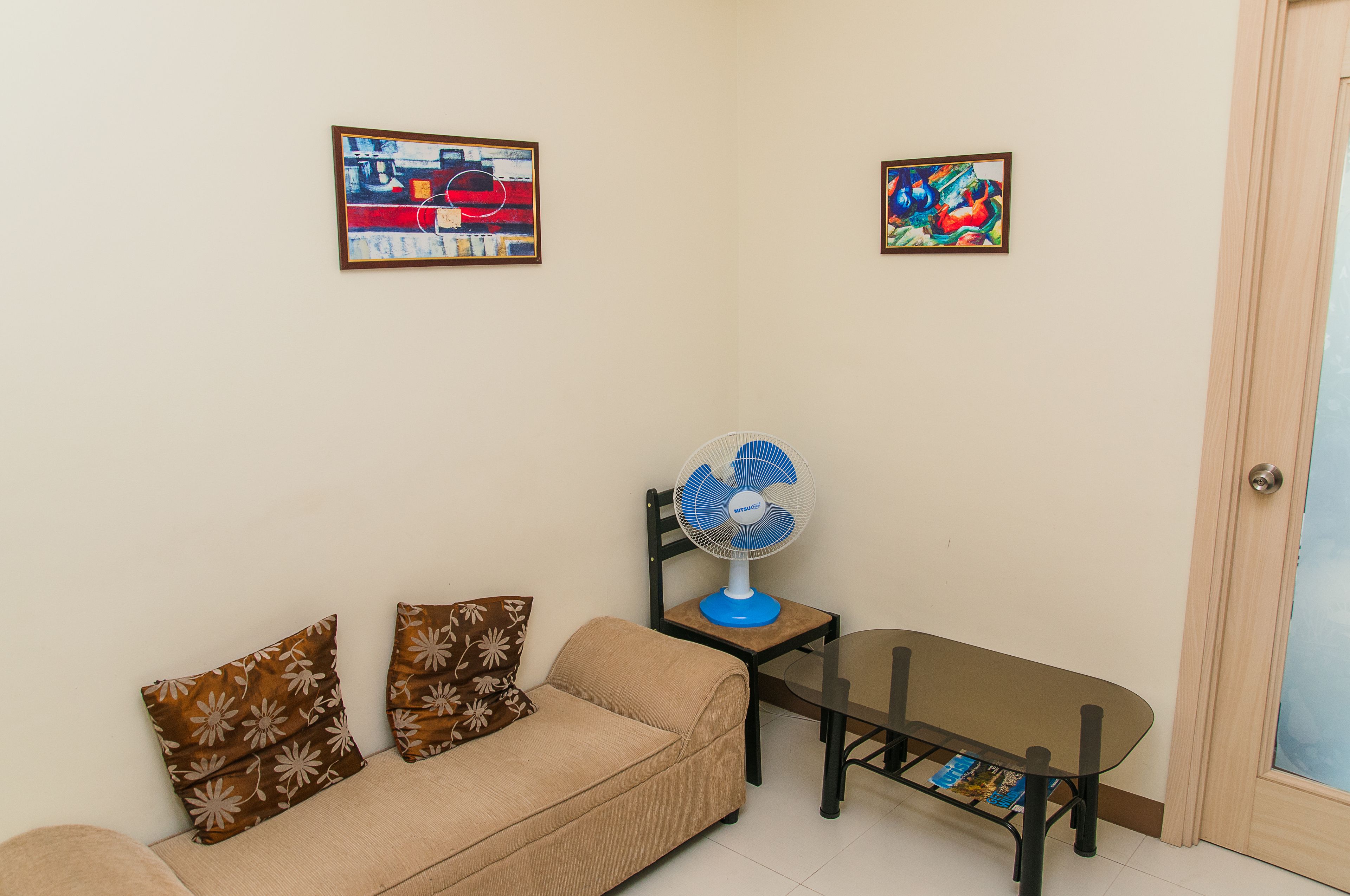 A living room with a sofa, a small table, a fan, and two framed artworks on the wall.