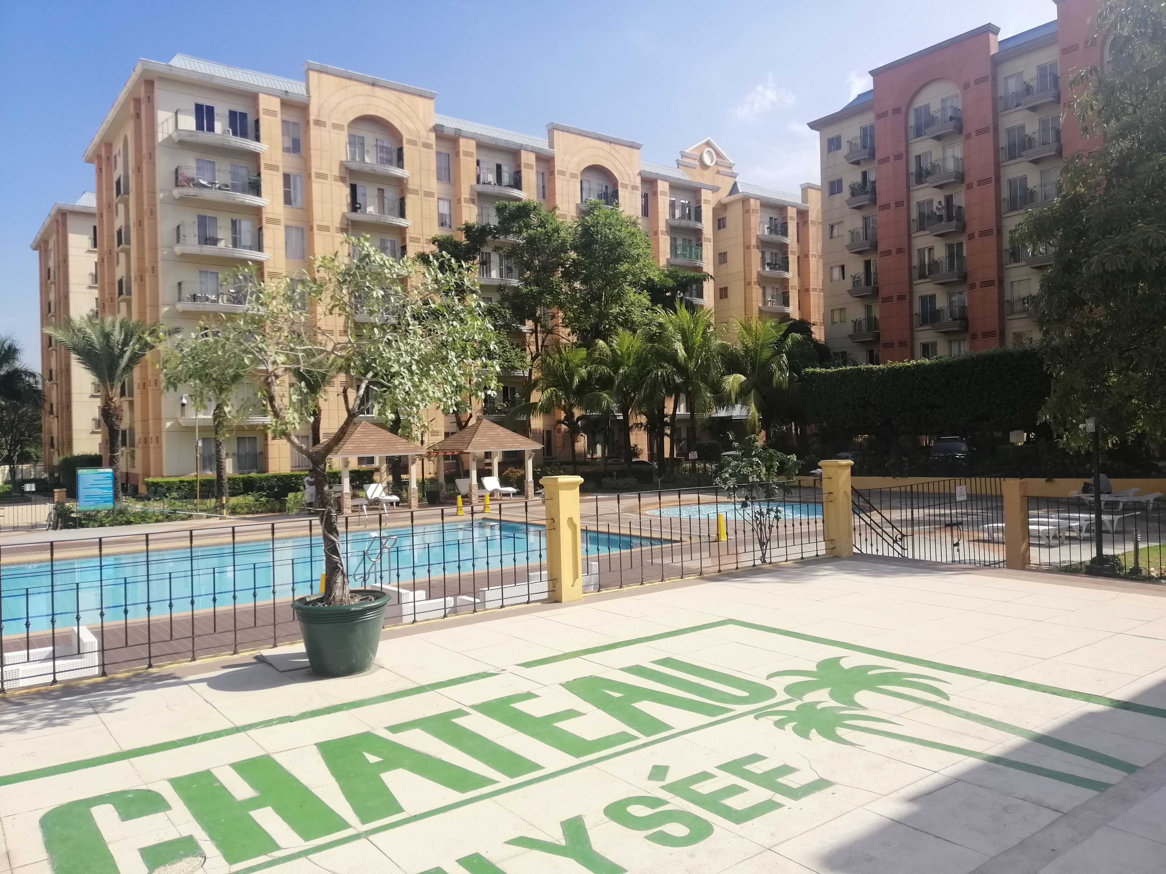 A hotel with a swimming pool, surrounded by palm trees and a fence.
