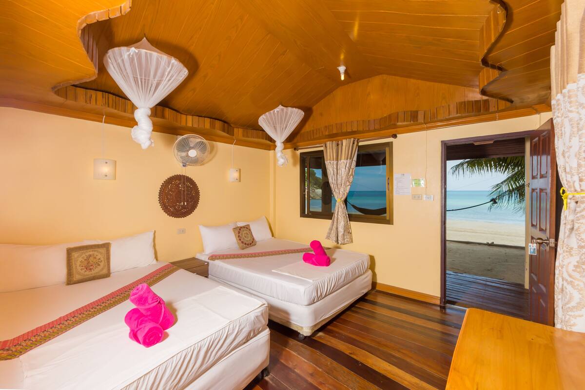A room with two beds, wooden flooring, a ceiling fan, a window with curtains, and a door leading to a beach view.