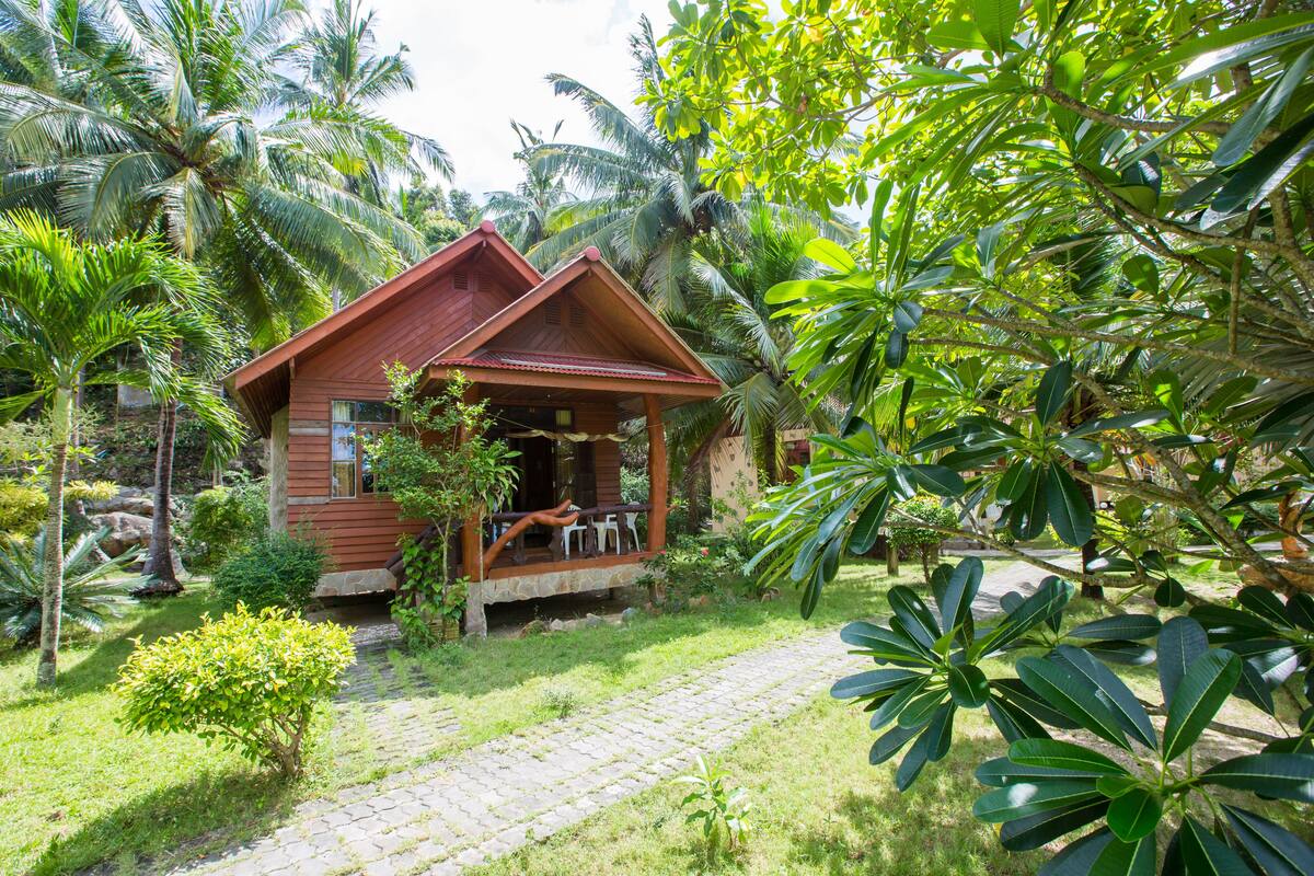 A wooden cabin with a porch, surrounded by lush greenery and palm trees.