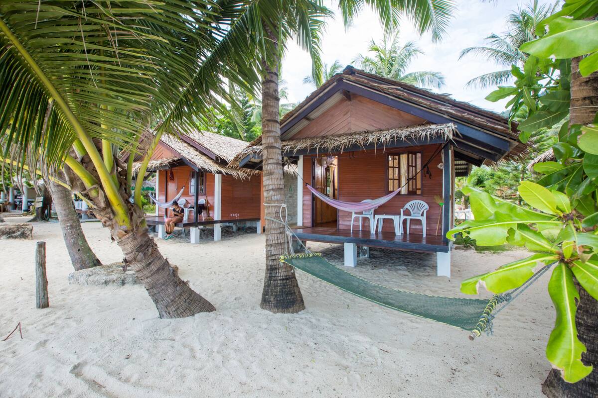 A beachside resort with wooden cabins and thatched roofs, surrounded by palm trees and a hammock.