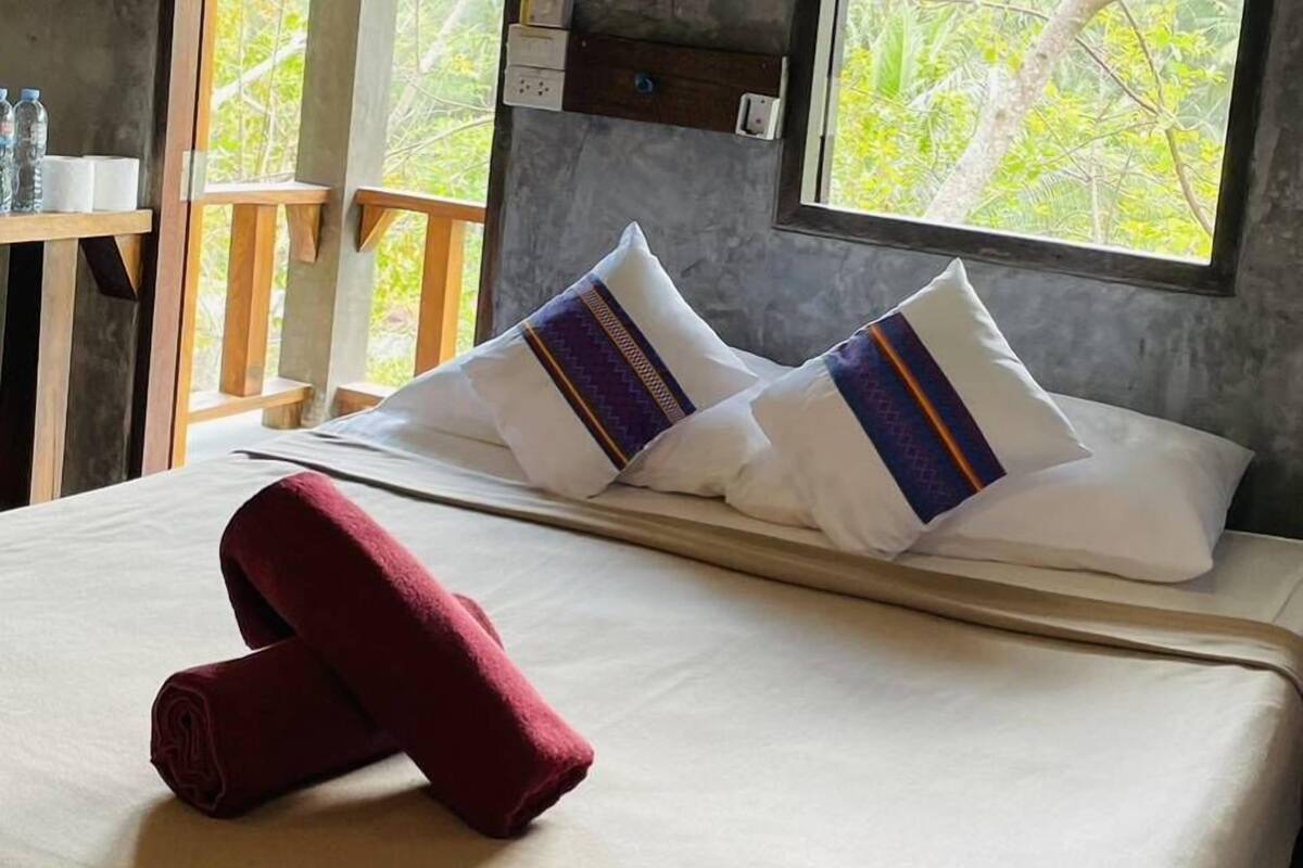 A hotel room with a bed, pillows, a red towel, a window with a view of trees, and a ceiling fan.