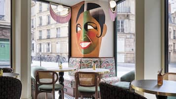 A café with round tables and wicker chairs, a large window with a view of buildings, and a colorful abstract painting on the wall.