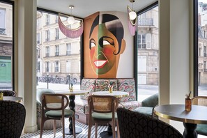 Un café avec des tables rondes et des chaises en osier, une grande fenêtre donnant sur des immeubles et une peinture abstraite aux couleurs vives accrochée au mur.