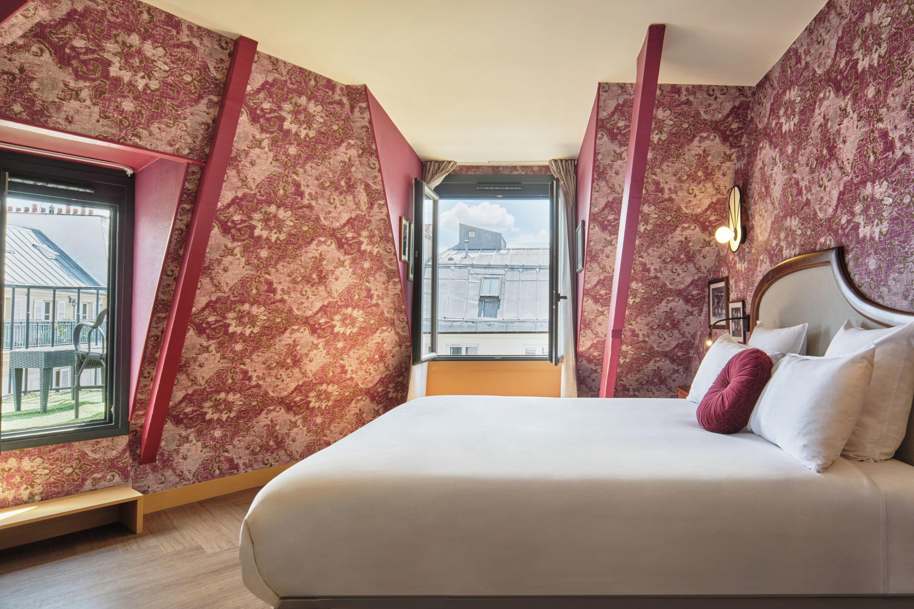 Ein Schlafzimmer mit einem großen Bett, floralem Wandpapier, einem Fenster mit Blick auf Gebäude und einem Nachttisch mit Lampe.