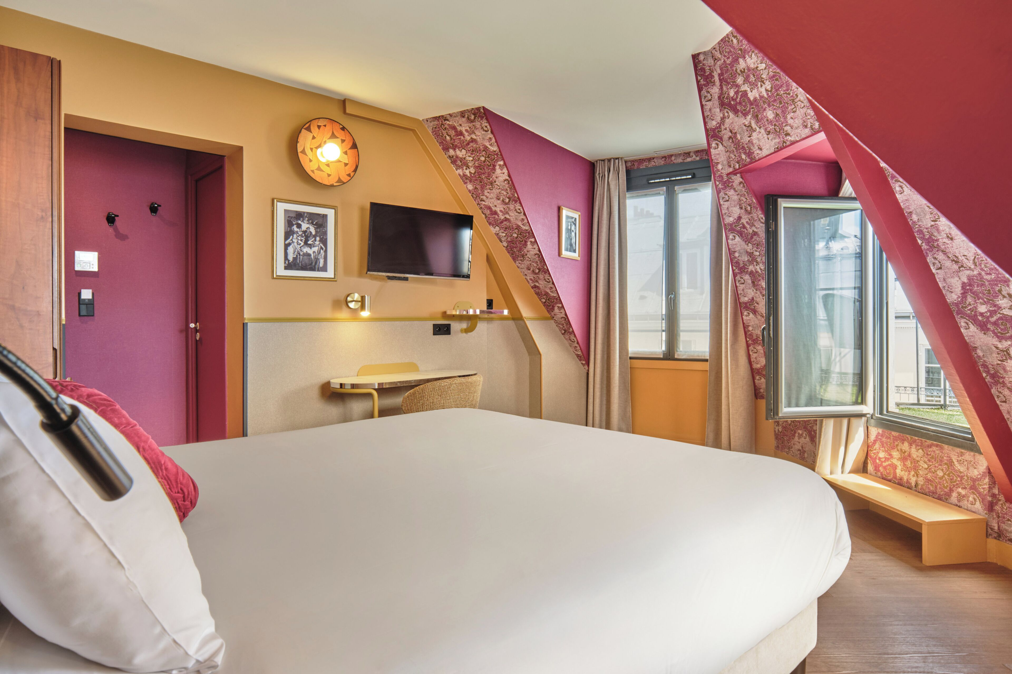 A hotel room with a large bed, a desk with a chair, a flat-screen TV mounted on the wall, and a window with a view of the outside.