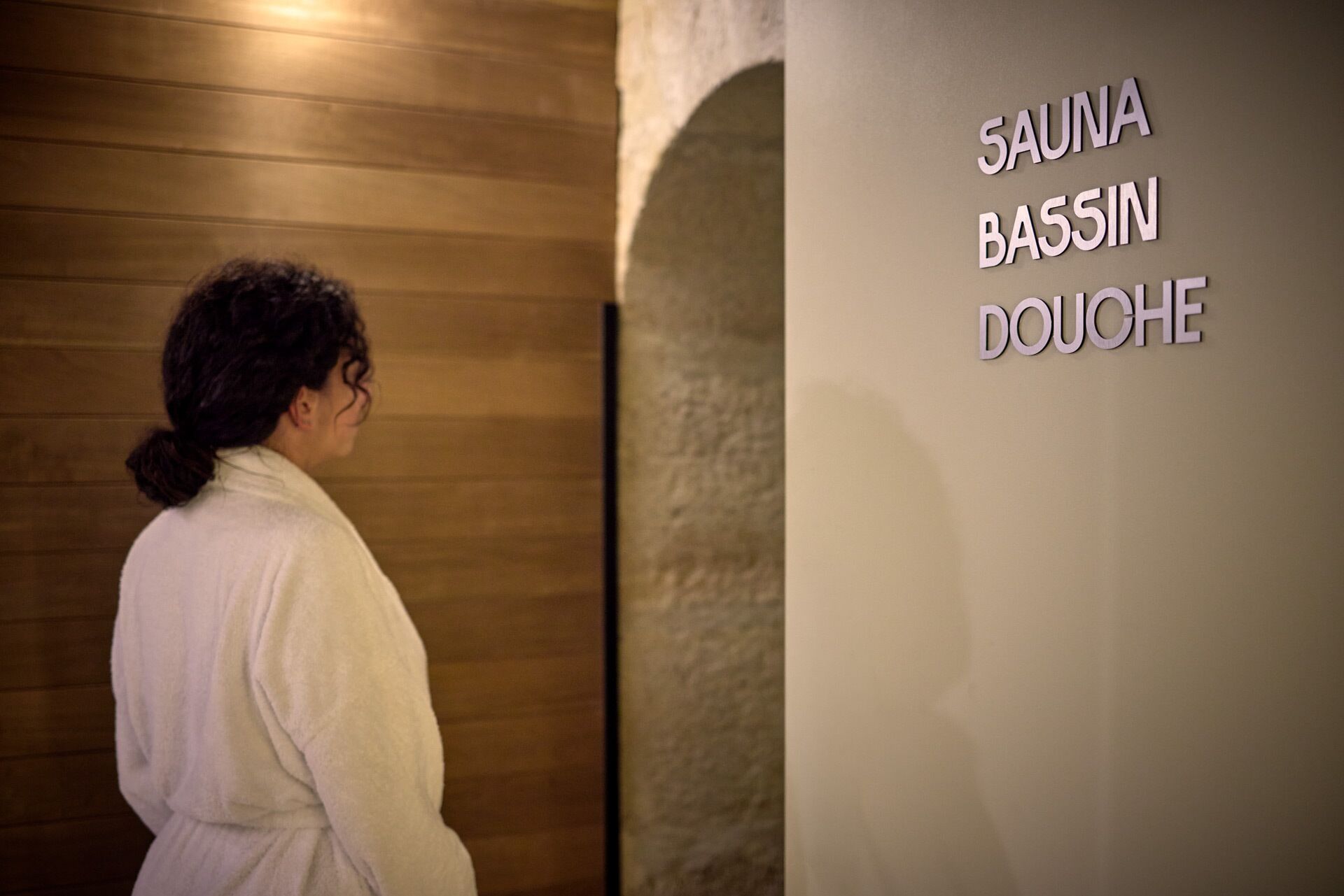 Una persona con un accappatoio bianco in prossimità di un muro con la scritta 'SAUNA BASSIN DOUCHE'.