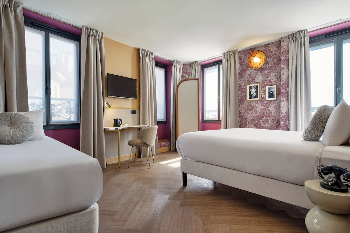 Habitación de hotel con dos camas, un escritorio, una silla, un televisor y un ventanal con cortinas.