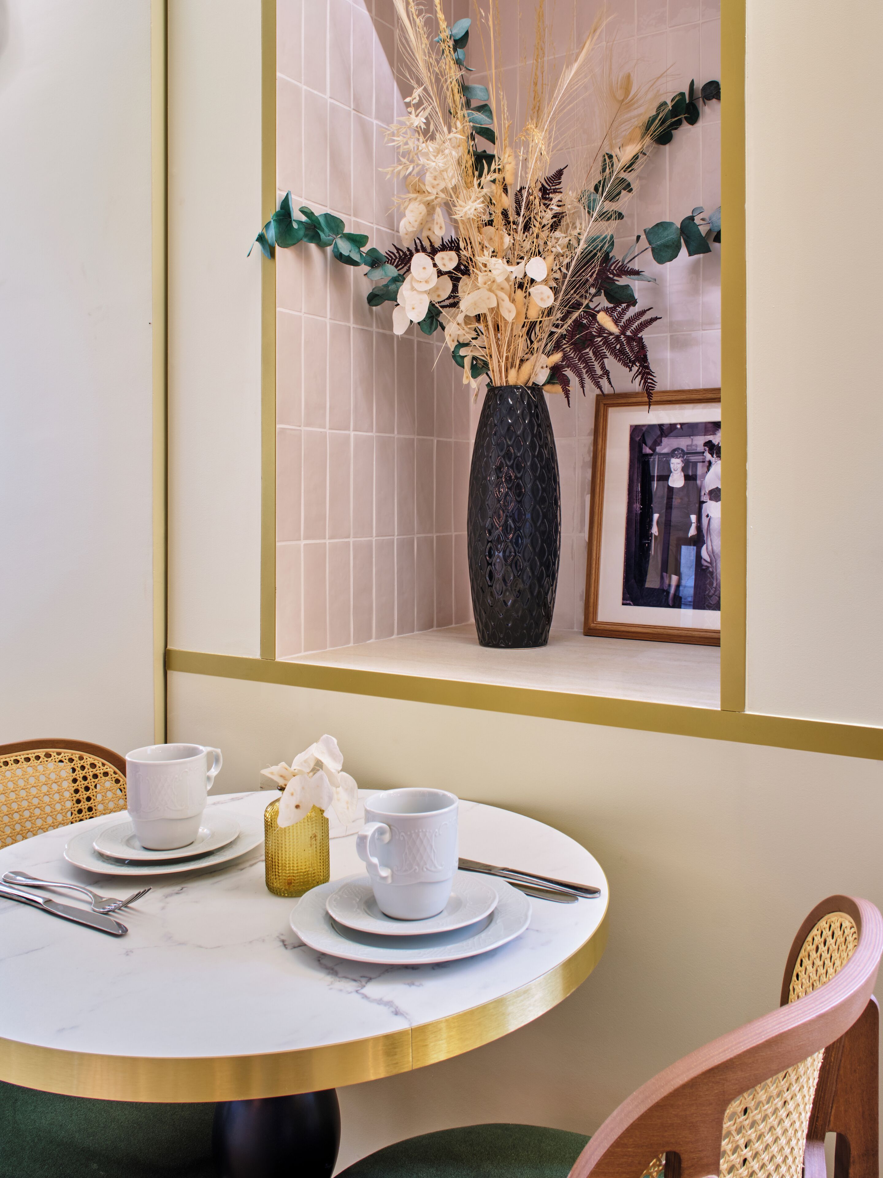 Une table de salle à manger ronde, avec de l’assiette et des tasses blanches, un vase contenant des plantes séchées et un tableau encadré posé sur une étagère.