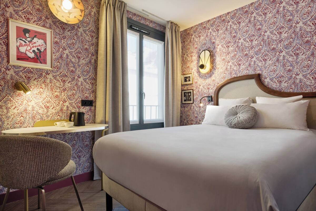 Habitación de hotel con una cama grande, un escritorio con silla, un cuadro en la pared y una ventana con cortinas.