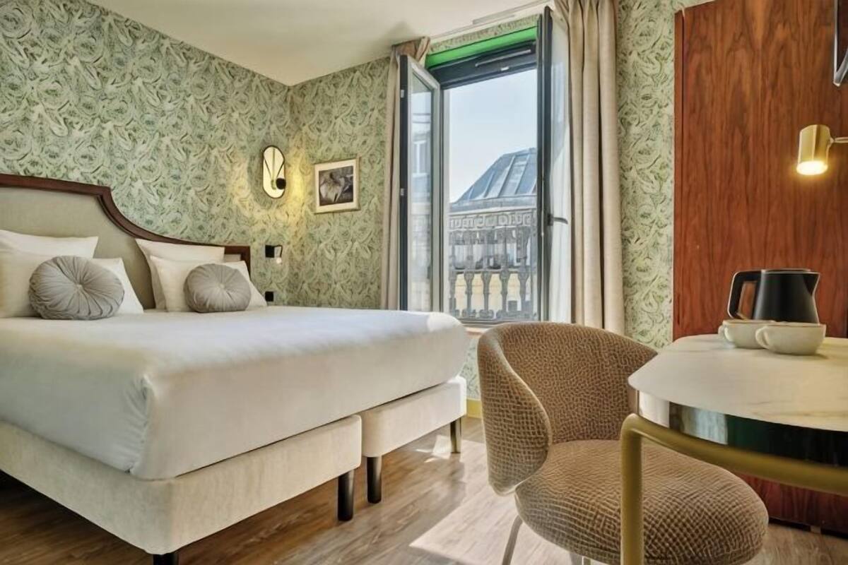 Habitación de hotel con cama, una silla, una mesa, un ventanal y un reloj y un cuadro en la pared.
