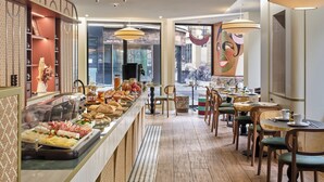 Un bufé de desayuno con diversas opciones de alimentos, suelo de madera y un área de estar con mesas y sillas.